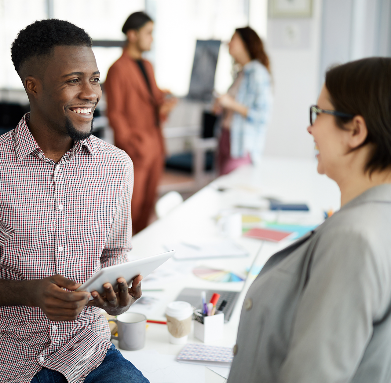 Two students standing near a desk in a modern office, engaged in conversation. One person is holding a tablet, while the desk displays papers, charts, and office supplies. In the background, other individuals are interacting near a whiteboard, creating a collaborative work environment.